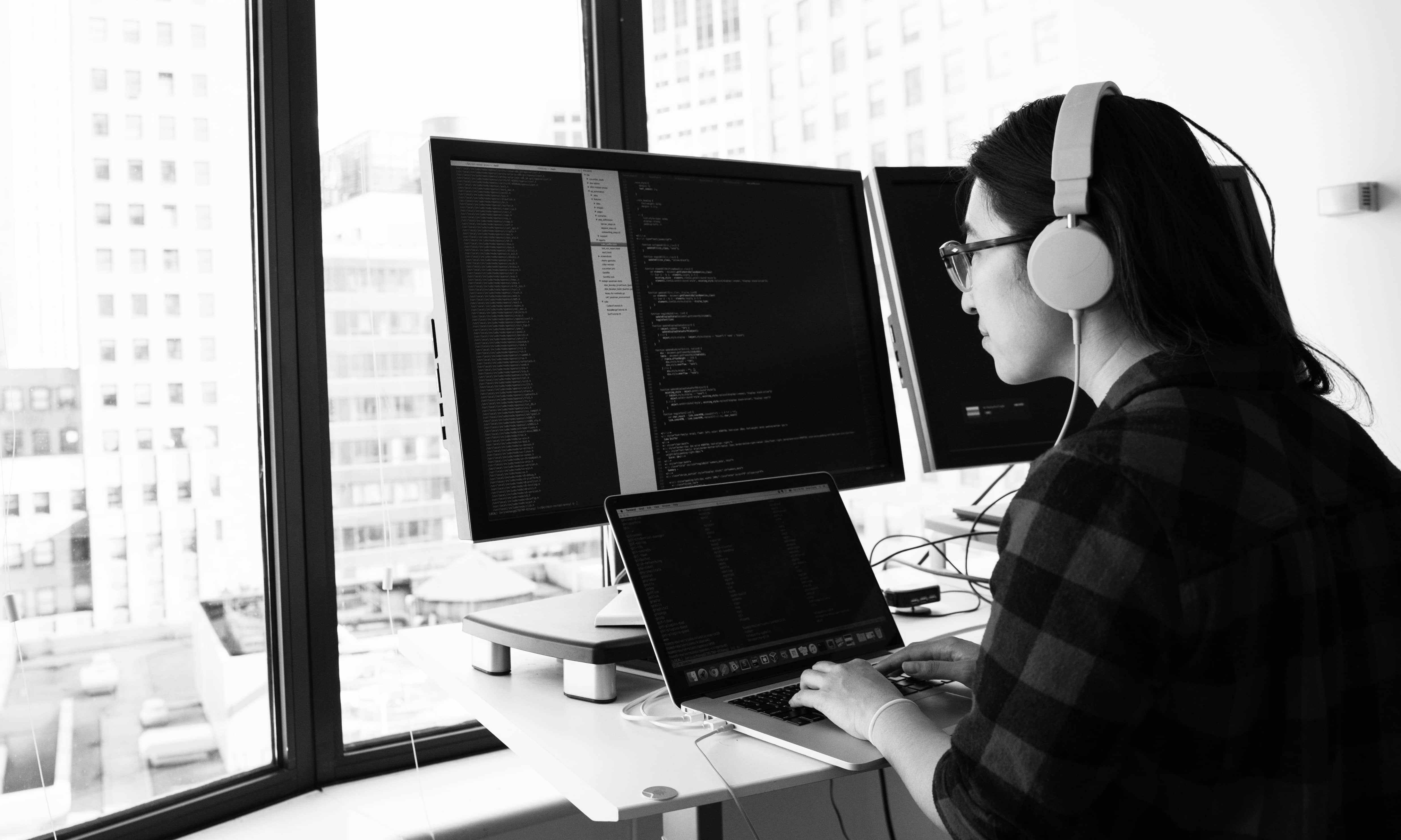 A black and white image of a female progammer in front of a big window wall coding with a laptop and two desktops.